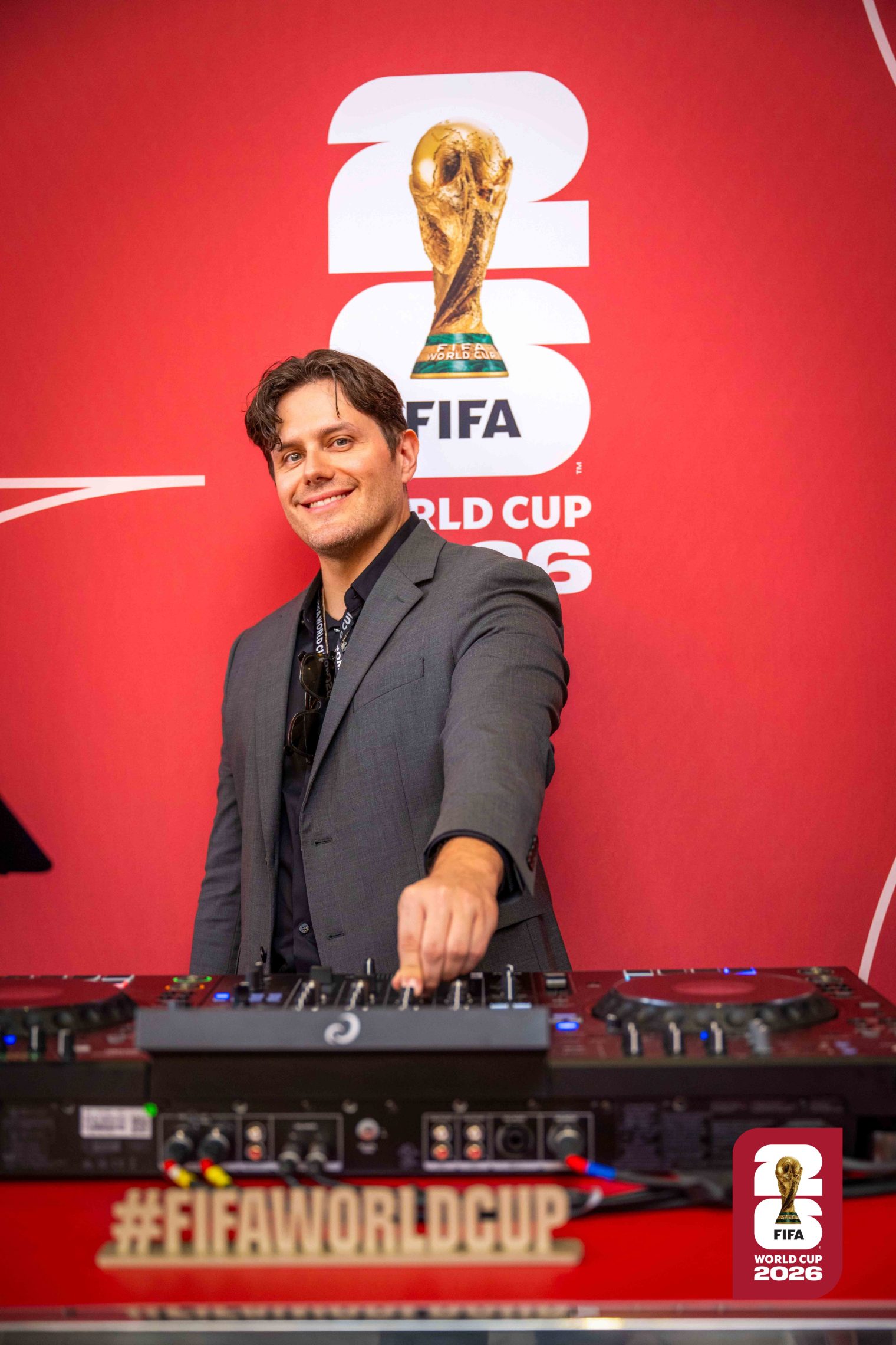 Elvis Suarez performing behind the decks, in a grey suit against a red event backdrop.